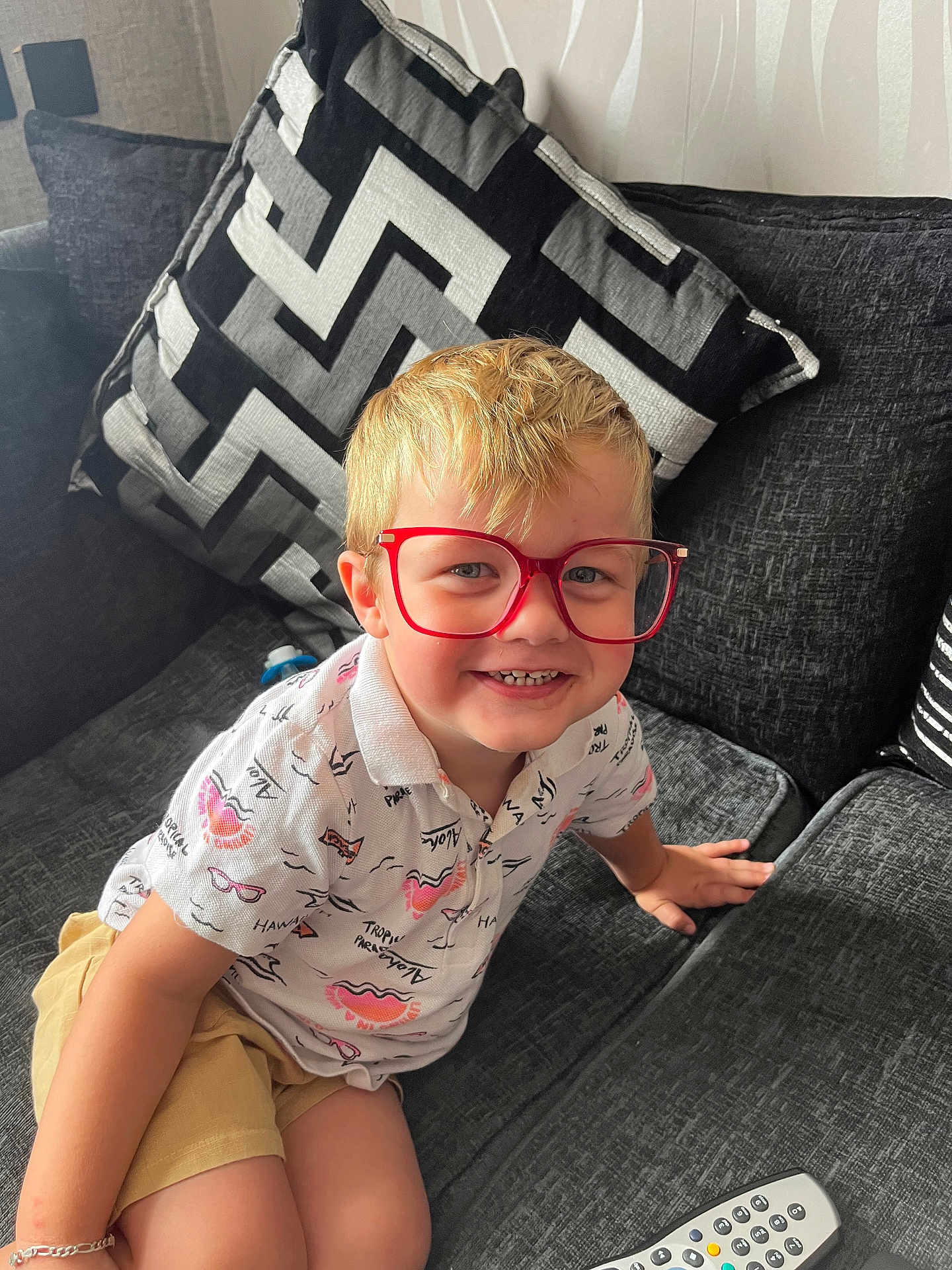 Leo is registered to the contest to win money with this photo: child, boy, glasses, red_glasses, smiling, couch, cushion, remote_control, shorts, shirt, indoor, blond_hair, happy, sitting, home, furniture, portrait, casual_clothing, playful, young