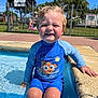 child, toddler, pool, water, swimsuit, sitting, smiling, sunny, outdoor, fence, sign, wet_hair, blue_clothing, playing, summer, happy, sunlight, concrete, recreation, vacation