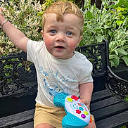 Leo is registered to the contest to win money with this photo: toddler, child, toy, game_controller, bench, outdoor, garden, plants, foliage, greenery, shorts, tshirt, blue_eyes, rosy_cheeks, curious, sitting, summer, daylight, person, playtime