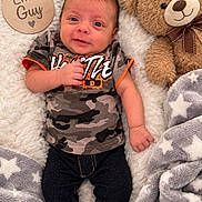 Jacob joined the competition — help win amazing prizes! baby, child, infant, portrait, smiling, lying_down, teddy_bear, stuffed_animal, blanket, fluffy_blanket, wooden_sign, text, camouflage_clothing, dark_pants, socks, nursery, indoor, close_up, happy, photography