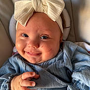 Darcie is registered to the contest to win money with this photo: baby, child, smiling, face, headband, bow, blue_clothing, hand, fingers, cute, infant, portrait, closeup, skin, happy, young, indoors, soft_light, expression, person