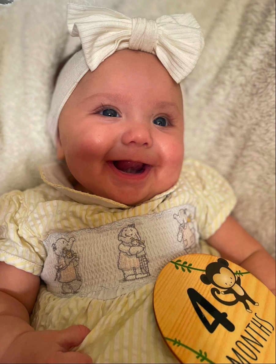 Darcie is registered to the contest to win money with this photo: baby, smiling, headband, bow, dress, yellow, bear_embroidery, wooden_sign, monkey, 4_months, milestone, child, happy, cute, portrait, infant, hand, skin, closeup, indoor