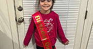 Selena is registered to the contest to win money with this photo: birthday, boots, casual_clothing, celebration, child, door, floor, hair, happy, holiday_theme, home, indoors, long_sleeve, person, red_sweater, sash, smile, standing, window_blinds, young
