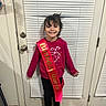 child, birthday, sash, smile, door, indoors, boots, long_sleeve, happy, standing, floor, window_blinds, red_sweater, person, young, celebration, hair, casual_clothing, holiday_theme, home