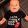 baby, child, infant, portrait, smiling, happy, sitting, leather_couch, black_clothing, onesie, rolling_stones, rock_and_roll, graphic_shirt, text_on_clothing, logo, indoors, close_up, face, hands, funny_expression