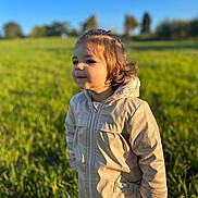 Soline a rejoint le concours — aidez-le/la à gagner de superbes lots ! toddler, child, jacket, beige, outdoor, field, grass, sunlight, blue_sky, curly_hair, portrait, person, nature, daylight, young_child, cute, happy, standing, clothing, warm_clothes