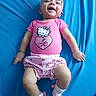 baby, pink_clothing, headband, bow, hello_kitty, smiling, lying_down, blue_background, socks, child, infant, cute, happy, playful, portrait, indoor, watermark, text_overlay, legs, arms