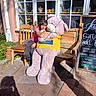 child, teddy_bear, hugging, smiling, outdoor, cafe, chalkboard, menu, wooden_bench, chair, window, shopfront, sunlight, brick_patio, flowers, potted_plants, pink_shirt, portrait, stuffed_animal, happy