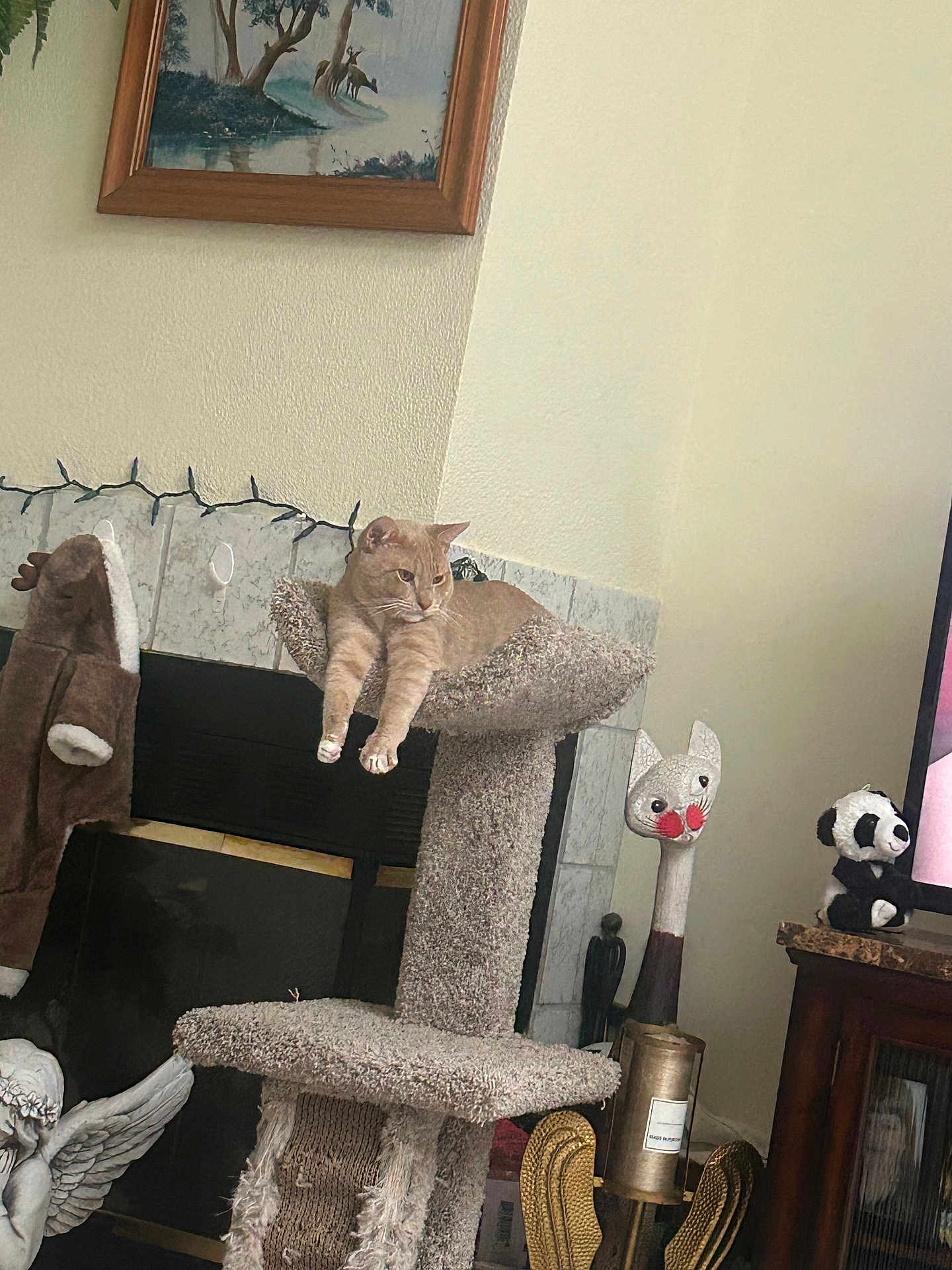 Simba is registered to the contest to win money with this photo: cat, cat_tree, fireplace, painting, stuffed_panda, wooden_cat_head, plush_toy, angel_statue, mantel, tile, living_room, decor, sitting, relaxed, whiskers, paws, scratching_post, television, cabinet, string_lights