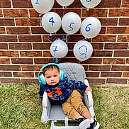 Advik is registered to the contest to win money with this photo: toddler, child, balloons, headphones, hoodie, orange_logo, chair, grass, brick_wall, outdoor, casual, shoes, baby, numbered_balloons, relaxed, portrait, cute, fashion, seated, playful