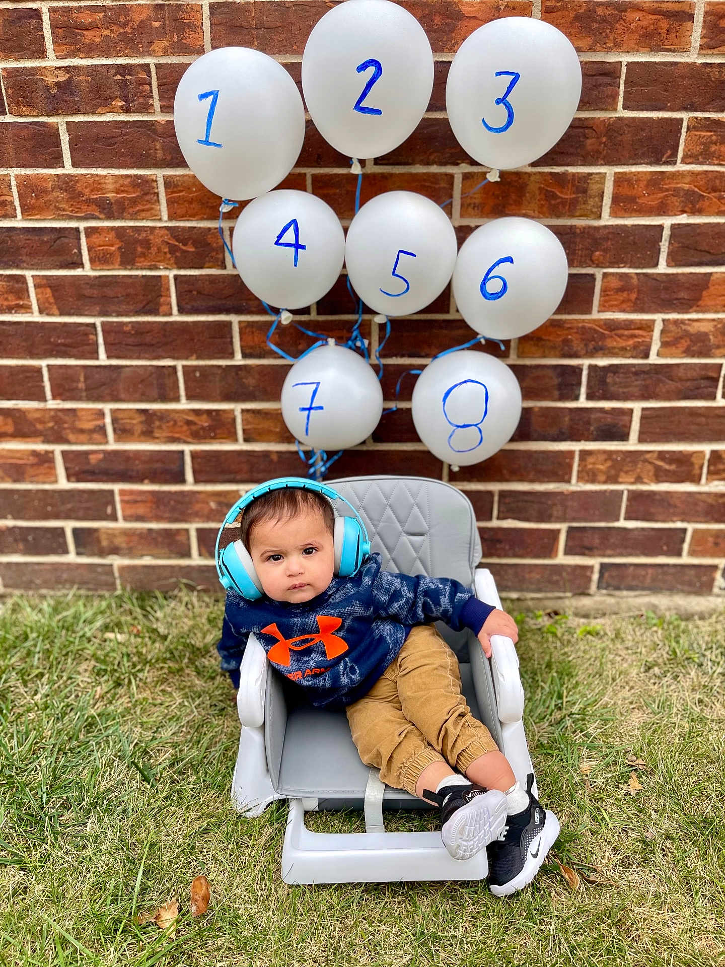 Advik is registered to the contest to win money with this photo: toddler, child, balloons, headphones, hoodie, orange_logo, chair, grass, brick_wall, outdoor, casual, shoes, baby, numbered_balloons, relaxed, portrait, cute, fashion, seated, playful