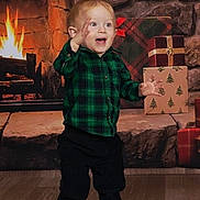 Phoenix is registered to the contest to win money with this photo: toddler, child, smiling, plaid_shirt, green_clothing, black_pants, standing, fireplace, fire, wrapped_gifts, holiday, indoor, wood_floor, stone_wall, excited, festive, cheerful, portrait, person, background