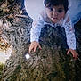 child, tree, bark, white_clothing, lace, outdoor, forest_floor, nature, curious, climbing, young_child, portrait, hands, face, eyes, daylight, texture, wood, person, innocence