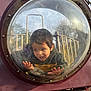 child, playground, plastic_dome, curious, hands, face, scratches, outdoor, park, play_equipment, metal_bars, hoodie, casual_clothing, daylight, trees, expression, young_child, peeking, fun, adventure