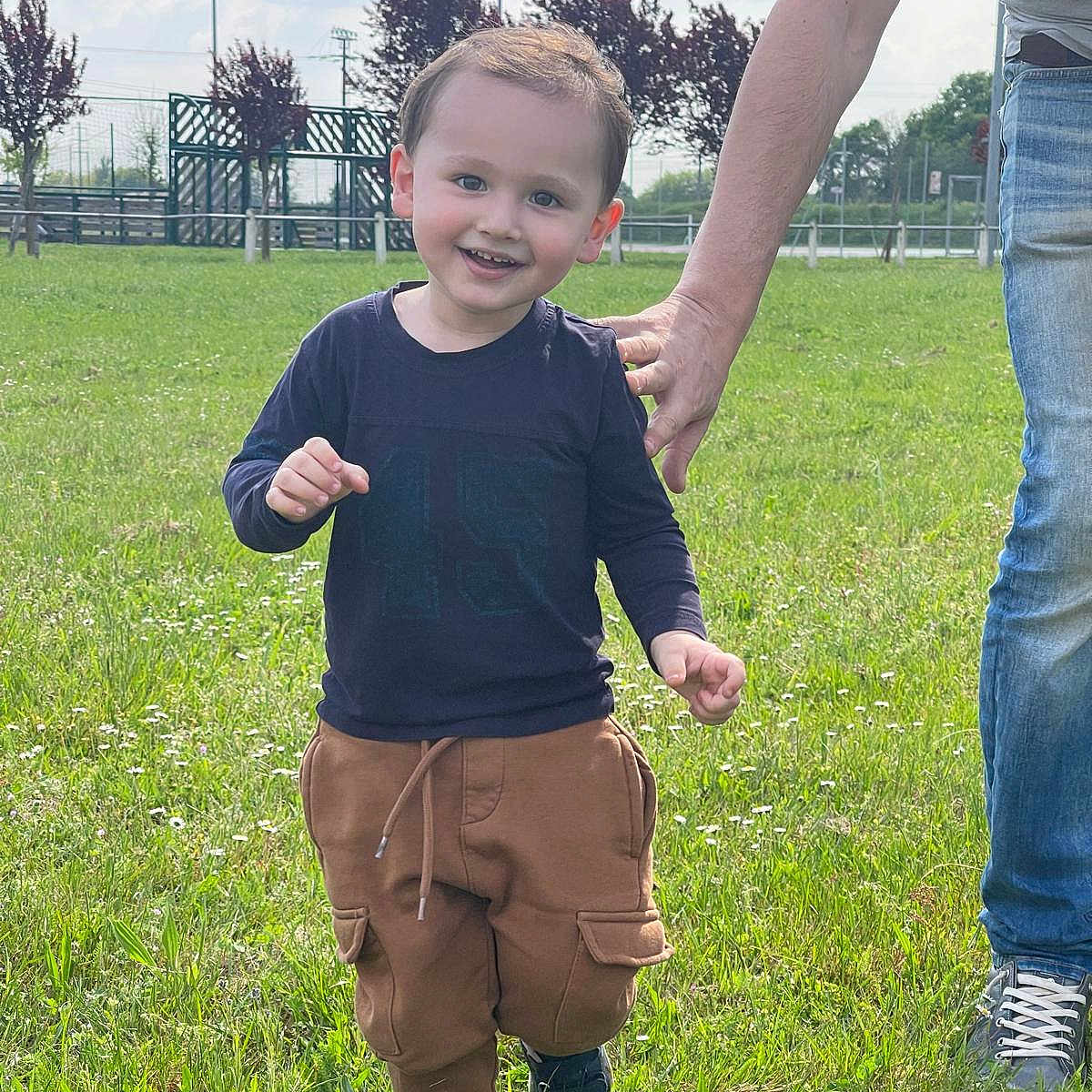 Ethan a rejoint le concours — aidez-le/la à gagner de superbes lots ! adult_hand, blue_shoes, boy, brown_pants, casual_clothing, child, daylight, field, grass, happy, long_sleeve_shirt, nature, outdoor, sky, smiling, sunny, support, toddler, trees, walking
