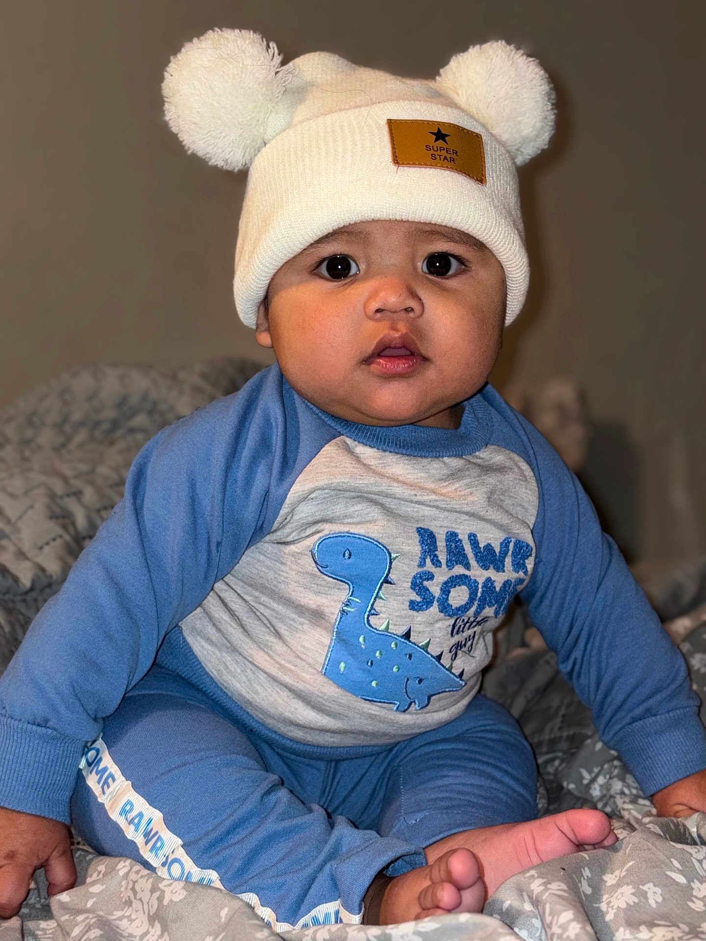 Dylan Nava joined the competition — help win amazing prizes! baby, child, hat, pom_poms, blue_clothing, dinosaur_graphic, sitting, blanket, indoors, cute, portrait, face, feet, hand, curious, wide_eyes, cozy, soft_lighting, infant, toddler