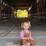 Finnleigh is registered to the contest to win money with this photo: child, girl, toddler, portrait, sitting, wooden_bridge, covered_bridge, outdoor, forest, sunlight, pigtails, pink_shirt, overalls, pink_shoes, road, wood, perspective, travel, photography, smiling