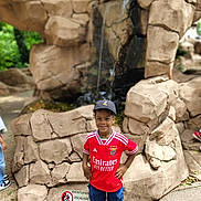 Ylan participe au concours pour gagner de l'argent avec cette photo : child, boy, smiling, red_jersey, cap, shorts, sneakers, waterfall, rocks, outdoor, daylight, nature, park, standing, confident, hands_on_hips, casual_clothing, happy, playful, scenery