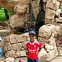 child, boy, smiling, red_jersey, cap, shorts, sneakers, waterfall, rocks, outdoor, daylight, nature, park, standing, confident, hands_on_hips, casual_clothing, happy, playful, scenery