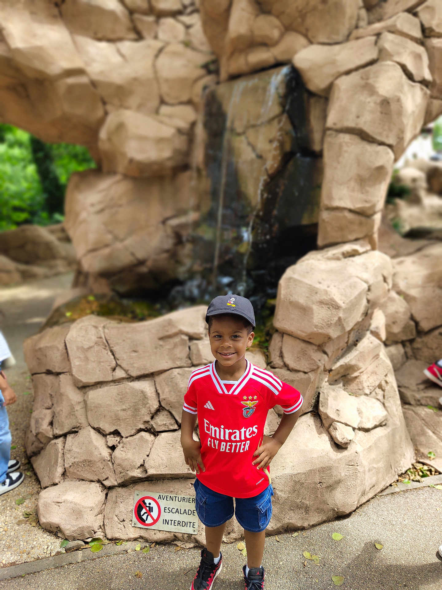 Ylan participe au concours pour gagner de l'argent avec cette photo : child, boy, smiling, red_jersey, cap, shorts, sneakers, waterfall, rocks, outdoor, daylight, nature, park, standing, confident, hands_on_hips, casual_clothing, happy, playful, scenery