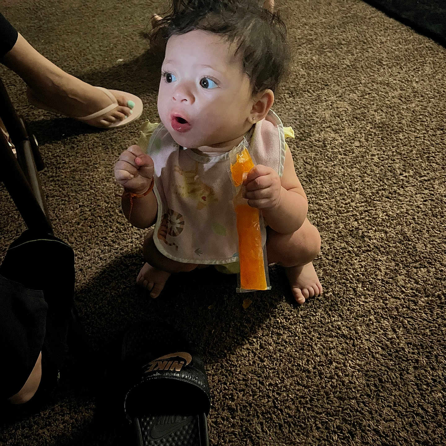 Makayla joined the competition — help win amazing prizes! baby, toddler, child, carpet, indoor, popsicle, food, footwear, sandal, slide, person, expression, curly_hair, bib, sitting, squatting, toy, background, light, shadow