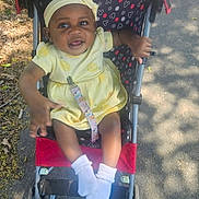 Ohana is registered to the contest to win money with this photo: baby, child, stroller, yellow_dress, headband, socks, outdoors, pavement, sunlight, shadow, leaf, happy, smiling, person, infant, cute, seat, pattern, hand, footwear