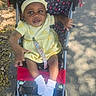 baby, child, stroller, yellow_dress, headband, socks, outdoors, pavement, sunlight, shadow, leaf, happy, smiling, person, infant, cute, seat, pattern, hand, footwear