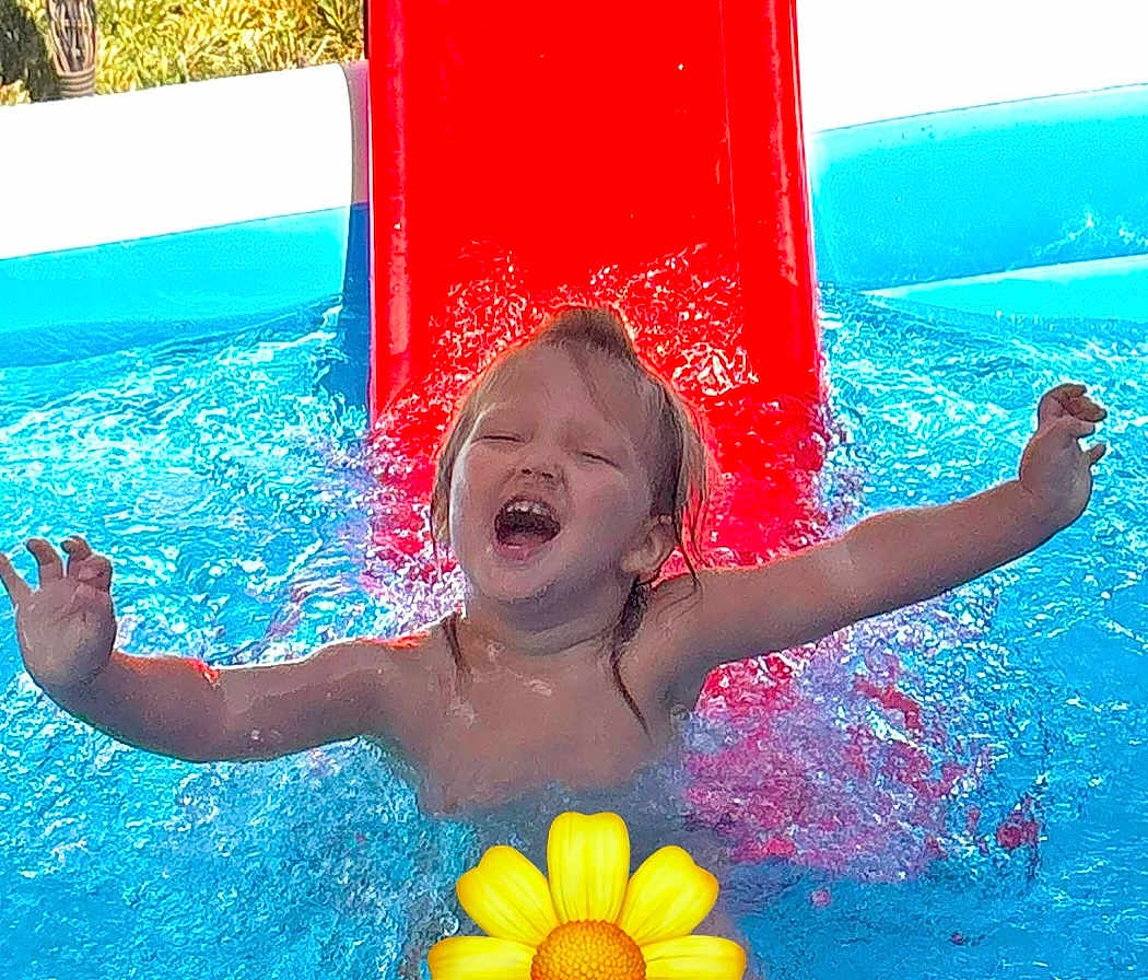 Lila is registered to the contest to win money with this photo: child, water, pool, slide, splash, happy, summer, outdoor, play, fun, yellow_flower_emoji, joy, swimming, arms_outstretched, daylight, wet_hair, blue_water, red_slide, person, smile