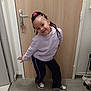 child, smile, pose, indoor, door, wooden_door, hoodie, braided_hair, pants, sneakers, floor, hand, happy, person, young, casual_clothing, standing, hairband, playful, home