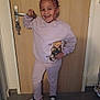 child, smiling, pose, indoors, door, sweatshirt, cat_design, pants, sneakers, floor, hair_braids, happy, casual_clothing, person, young, standing, bracelet, keychain, wooden_door, home