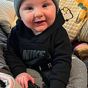 Louis joined the competition — help win amazing prizes! baby, beanie, blanket, child, clothing, cozy, cushion, cute, gray_pants, home, hoodie, indoors, infant, nike, person, remote_control, sitting, smiling, toy, white_socks