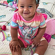 Sayge is registered to the contest to win money with this photo: baby, child, smiling, portrait, sitting, indoor, play_mat, unicorn, toy, headband, flower, curly_hair, pink_clothing, rainbow, onesie, shoes, infant, happy, playroom, close_up