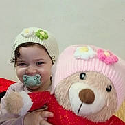 Dilara participe au concours pour gagner de l'argent avec cette photo : child, cozy, curly_hair, cute, flower_decoration, happy, hat, holding, indoor, knit_hat, pacifier, person, pink_hat, plush_toy, portrait, red_sweater, smile, soft_toy, teddy_bear, toddler