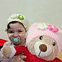 child, cozy, curly_hair, cute, flower_decoration, happy, hat, holding, indoor, knit_hat, pacifier, person, pink_hat, plush_toy, portrait, red_sweater, smile, soft_toy, teddy_bear, toddler