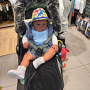 Syreese is registered to the contest to win money with this photo: baby, stroller, hat, blue_bib, toy, person, shopping, store, clothing, floor, footwear, hand, jacket, pants, socks, shoes, fur_cover, curious, indoor, child