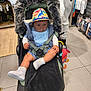 baby, stroller, hat, blue_bib, toy, person, shopping, store, clothing, floor, footwear, hand, jacket, pants, socks, shoes, fur_cover, curious, indoor, child
