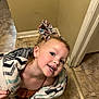 child, smiling, blanket, bow, hair_accessory, indoor, floor, tile_floor, person, happy, cute, young_girl, earrings, home, wall, doorframe, playful, portrait, clothing, head