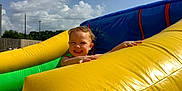 Hazel joined the competition — help win amazing prizes! child, toddler, smiling, inflatable, yellow, blue, green, outdoor, playground, slide, summer, sunny, happy, fun, sky, clouds, fence, play, person, recreation