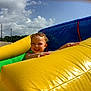 child, toddler, smiling, inflatable, yellow, blue, green, outdoor, playground, slide, summer, sunny, happy, fun, sky, clouds, fence, play, person, recreation