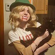 Michael is registered to the contest to win money with this photo: person, cat, black_cat, costume, wig, green_hat, fake_blood, fangs, indoor, wooden_cabinet, bracelet, long_sleeve_shirt, tshirt, blonde_hair, holding, pet, face, expression, home, casual