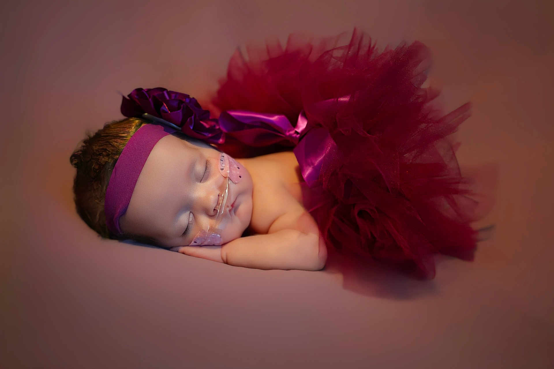 Alexandria is registered to the contest to win money with this photo: baby, sleeping, tutu, headband, flower, nasal_cannula, medical_tape, infant, portrait, soft_lighting, maroon, peaceful, resting, newborn, cute, closeup, child, studio_photo, pink_background, adorable