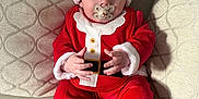 Hudson joined the competition — help win amazing prizes! baby, santa_claus_costume, pacifier, red_clothing, hat, couch, cushion, indoor, child, cute, holiday, christmas, plush_slippers, reindeer_slippers, infant, sitting, person, face, hands, cozy