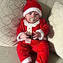 baby, santa_claus_costume, pacifier, red_clothing, hat, couch, cushion, indoor, child, cute, holiday, christmas, plush_slippers, reindeer_slippers, infant, sitting, person, face, hands, cozy