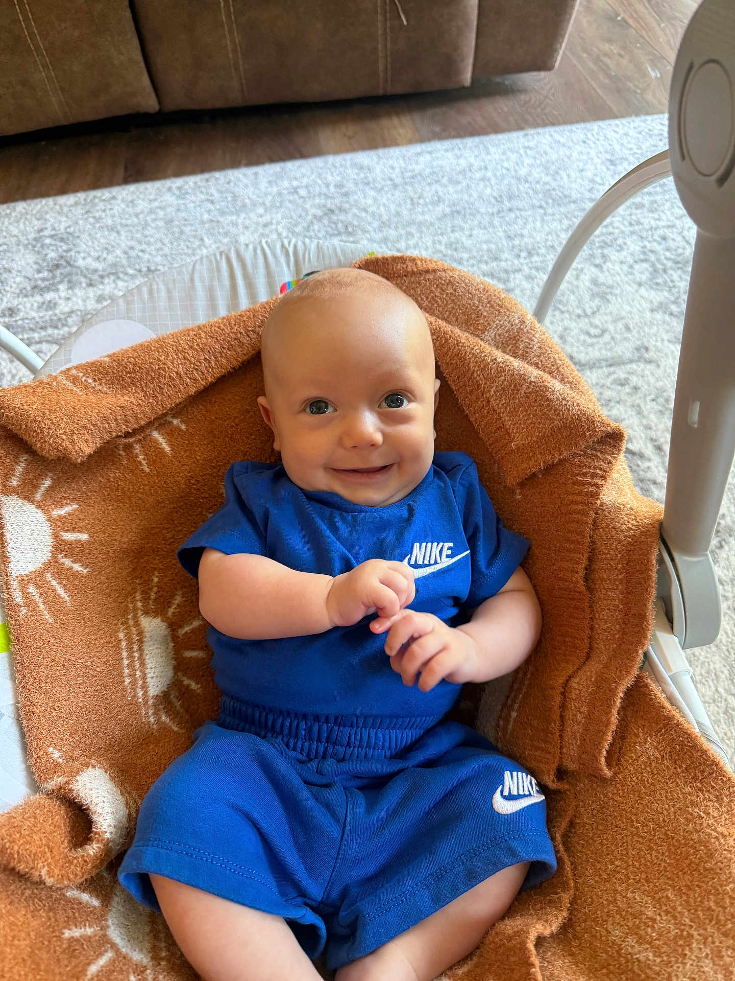 Silas is registered to the contest to win money with this photo: baby, smiling, blue_clothing, nike, blanket, brown_blanket, sun_pattern, infant, happy, cute, indoor, carpet, furniture, baby_bouncer, child, person, portrait, sitting, small_hands, head