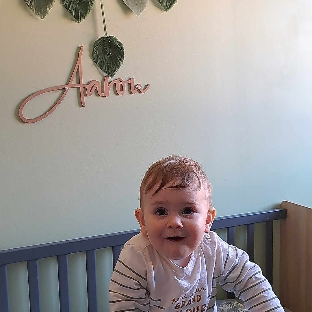 Aaron a rejoint le concours — aidez-le/la à gagner de superbes lots ! toddler, crib, child, baby, nursery, decor, wall, hanging_leaves, name_sign, furniture, indoors, smiling, curious, person, bed, cot, clothing, striped_shirt, hand, standing