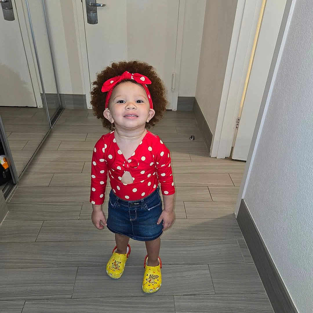 Zayah joined the competition — help win amazing prizes! child, clothing, curly_hair, cute, denim_skirt, door, flooring, footwear, hallway, happy, indoor, mirror, person, polka_dot, portrait, red_headband, smiling, standing, toddler, yellow_clogs