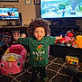 Zayah joined the competition — help win amazing prizes! carpet, child, clothing, curly_hair, entertainment, footwear, furniture, green_shirt, home, indoor, living_room, monitor, person, photo_frames, pink_toy_car, plush_toys, smiling, television, toddler, toys