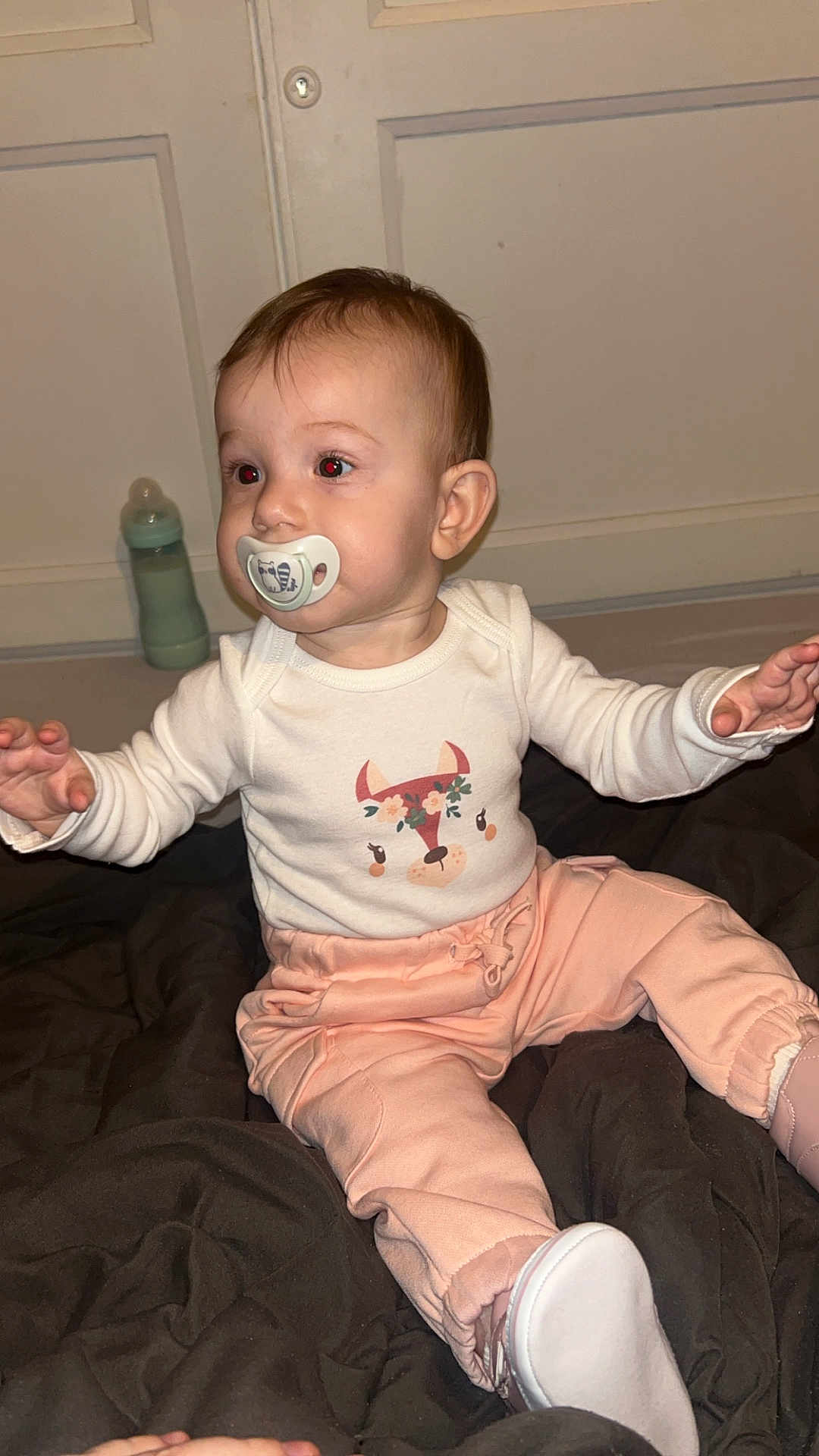Ilyana a rejoint le concours — aidez-le/la à gagner de superbes lots ! baby, child, pacifier, infant, sitting, white_shirt, peach_pants, footwear, blanket, indoors, cute, clothing, face, expression, hand, person, young_child, room, curious, floor