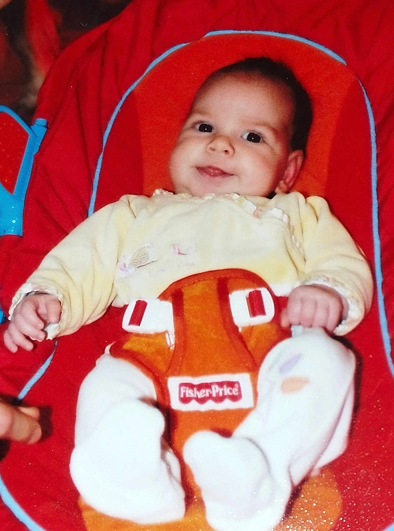 Lilie participe au concours pour gagner de l'argent avec cette photo : baby, infant, smile, fisher_price, baby_seat, red, orange, yellow_clothing, white_socks, hand, face, child, cute, indoor, toy, seat_belt, plush, comfort, young_child, portrait