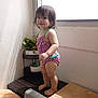 toddler, child, swimsuit, standing, indoor, window, sunlight, plant, greenery, wooden_surface, barefoot, hair, face, smile, corner, shadow, bright, colorful, room, casual
